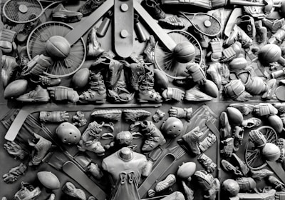 A complex arrangement of various sports equipment and attire, including tennis rackets, soccer balls, basketballs, helmets, shoes, gloves, and a mannequin torso wearing a T-shirt. The items are layered and artistically displayed in a monochrome color scheme, creating an intricate texture and visual pattern.