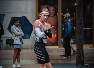 A person wearing a sporty outfit, including a headband, tight-fitting tank top, and shorts, is walking on a street. They appear to be gesturing playfully with their hands. In the background, a store with advertisements, and people, some of whom are holding umbrellas, are visible standing on the sidewalk.