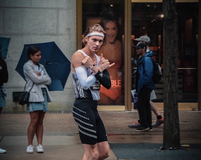 A person wearing a sporty outfit, including a headband, tight-fitting tank top, and shorts, is walking on a street. They appear to be gesturing playfully with their hands. In the background, a store with advertisements, and people, some of whom are holding umbrellas, are visible standing on the sidewalk.