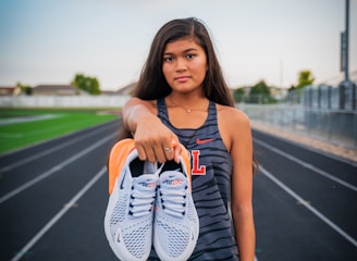 A young woman stands on a running track, holding a pair of white sneakers with orange details directly towards the viewer. She wears athletic clothing, including a sleeveless top with a letter 'L' on it and black shorts. The background features a sports field and a cloudy sky, suggesting a typical outdoor sports environment.
