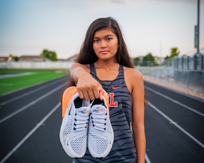 A young woman stands on a running track, holding a pair of white sneakers with orange details directly towards the viewer. She wears athletic clothing, including a sleeveless top with a letter 'L' on it and black shorts. The background features a sports field and a cloudy sky, suggesting a typical outdoor sports environment.