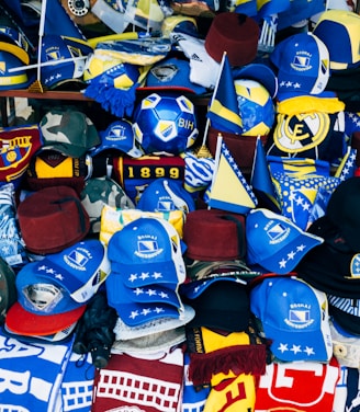 A vibrant display of sports merchandise with various hats, caps, and flags in blue and yellow, prominently featuring the Bosnia and Herzegovina flag and football-related items. There are shirts and scarves with team logos, including a jersey with the name Mbappe and the number 29.