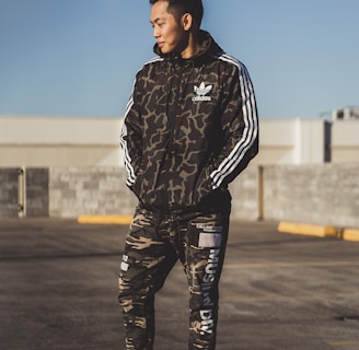 A person stands outdoors wearing a camouflage-patterned athletic outfit with the logo of a well-known sports brand. The individual appears to be in a parking lot or open area under clear daylight. The outfit consists of a jacket with white stripes on the sleeves and matching pants, paired with white sneakers.