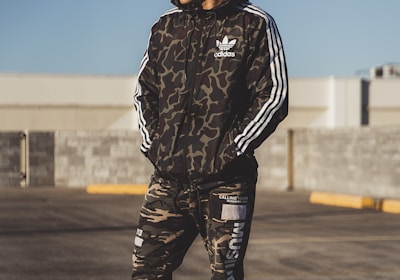 A person stands outdoors wearing a camouflage-patterned athletic outfit with the logo of a well-known sports brand. The individual appears to be in a parking lot or open area under clear daylight. The outfit consists of a jacket with white stripes on the sleeves and matching pants, paired with white sneakers.