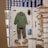 A retail display inside a store featuring outdoor or casual clothing and accessories arranged on a white board. Items include a green long-sleeve shirt, gray pants, a pair of green and beige shoes, a green hat, sunglasses, a pair of white socks, a small green item, and a blue pouch. In the background, shelves are stacked with blue and white shoe boxes.