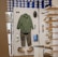 A retail display inside a store featuring outdoor or casual clothing and accessories arranged on a white board. Items include a green long-sleeve shirt, gray pants, a pair of green and beige shoes, a green hat, sunglasses, a pair of white socks, a small green item, and a blue pouch. In the background, shelves are stacked with blue and white shoe boxes.