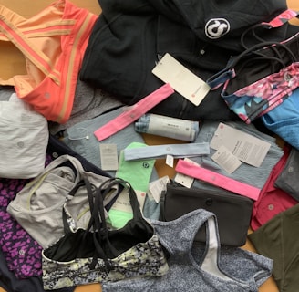A collection of various athletic apparel and accessories laid out, including sports bras, tops, headbands, and tags. The items are in different colors and patterns, such as pastel pink, blue, and floral designs. A black jacket and several pieces still have brand tags attached.