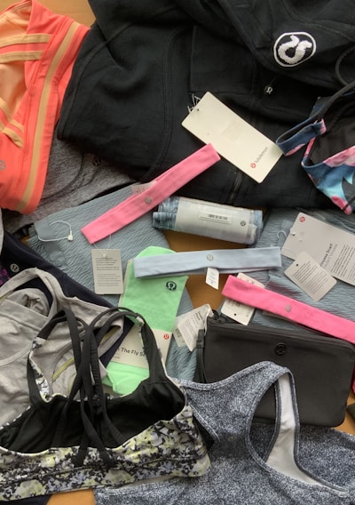 A collection of various athletic apparel and accessories laid out, including sports bras, tops, headbands, and tags. The items are in different colors and patterns, such as pastel pink, blue, and floral designs. A black jacket and several pieces still have brand tags attached.