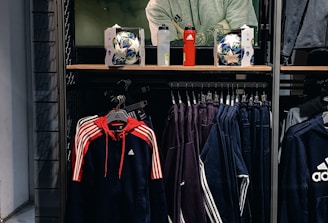 A store display features a large screen showcasing a man in a grey hoodie with the Adidas logo, while below it are two Adidas soccer balls and a red bottle. The clothing rack beneath holds hanging Adidas jackets, including a prominent navy blue and red jacket with white stripes, alongside dark-colored track pants.