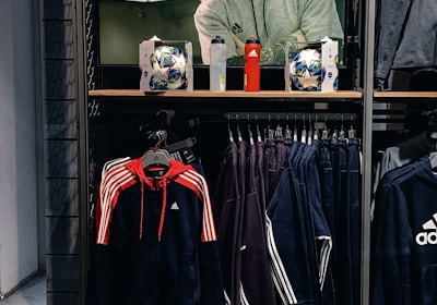 A store display features a large screen showcasing a man in a grey hoodie with the Adidas logo, while below it are two Adidas soccer balls and a red bottle. The clothing rack beneath holds hanging Adidas jackets, including a prominent navy blue and red jacket with white stripes, alongside dark-colored track pants.