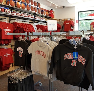 A retail display in a store features Stanford University merchandise, including red and grey t-shirts, hoodies, and sports equipment like basketballs and footballs on shelves and racks.