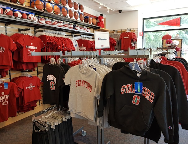 A retail display in a store features Stanford University merchandise, including red and grey t-shirts, hoodies, and sports equipment like basketballs and footballs on shelves and racks.