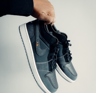 A hand is holding a pair of dark-colored athletic shoes. The shoes have a combination of leather and mesh materials with a swoosh logo on the side. They feature black laces and white soles. The hand appears to be wearing a black sleeve.