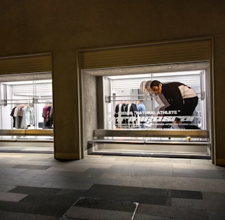 A store display with two large glass windows, featuring clothing racks with various items such as shirts and jackets. There is a poster of a person bending down, with text 'Natural Athlete' across it. The surrounding area includes a tiled walkway.