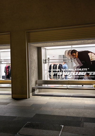 A store display with two large glass windows, featuring clothing racks with various items such as shirts and jackets. There is a poster of a person bending down, with text 'Natural Athlete' across it. The surrounding area includes a tiled walkway.