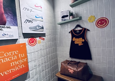 A small retro-themed display with a focus on athletic apparel and shoes. The walls feature a series of posters showcasing various Nike shoes from different years. A black gym tank top and a brown leather gym bag are prominently positioned on a wooden bench. There are decorative elements like potted plants on wall-mounted shelves and smiley face icons. The environment has a nostalgic and sporty atmosphere.