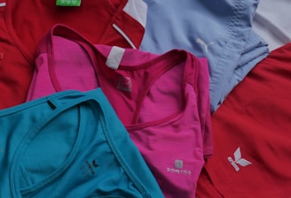 Several sports tank tops in an assortment of colors including red, pink, blue, and turquoise, arranged overlapping one another. The tops feature logos from different brands.