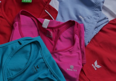 Several sports tank tops in an assortment of colors including red, pink, blue, and turquoise, arranged overlapping one another. The tops feature logos from different brands.