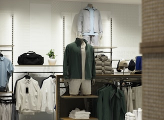 A retail clothing store display featuring mannequins dressed in casual, sporty attire including jackets and shorts. Shelves and racks are organized with neatly folded shirts and jackets in neutral and earthy tones. The lighting is bright and modern, enhancing the sleek display of garments and accessories.