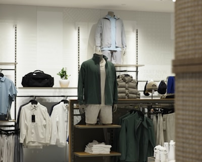 A retail clothing store display featuring mannequins dressed in casual, sporty attire including jackets and shorts. Shelves and racks are organized with neatly folded shirts and jackets in neutral and earthy tones. The lighting is bright and modern, enhancing the sleek display of garments and accessories.